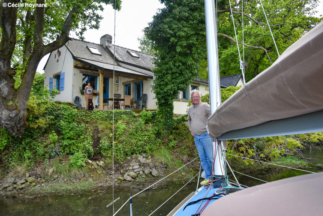 Portrait environnemental du constructeurs de voiliers catboat, Antoile Carmichaël, qui pose à l'avant de son bateau sur la rivière de l'Odet que sa maison surplombe. Un décor de forêt typique de Bretagne.